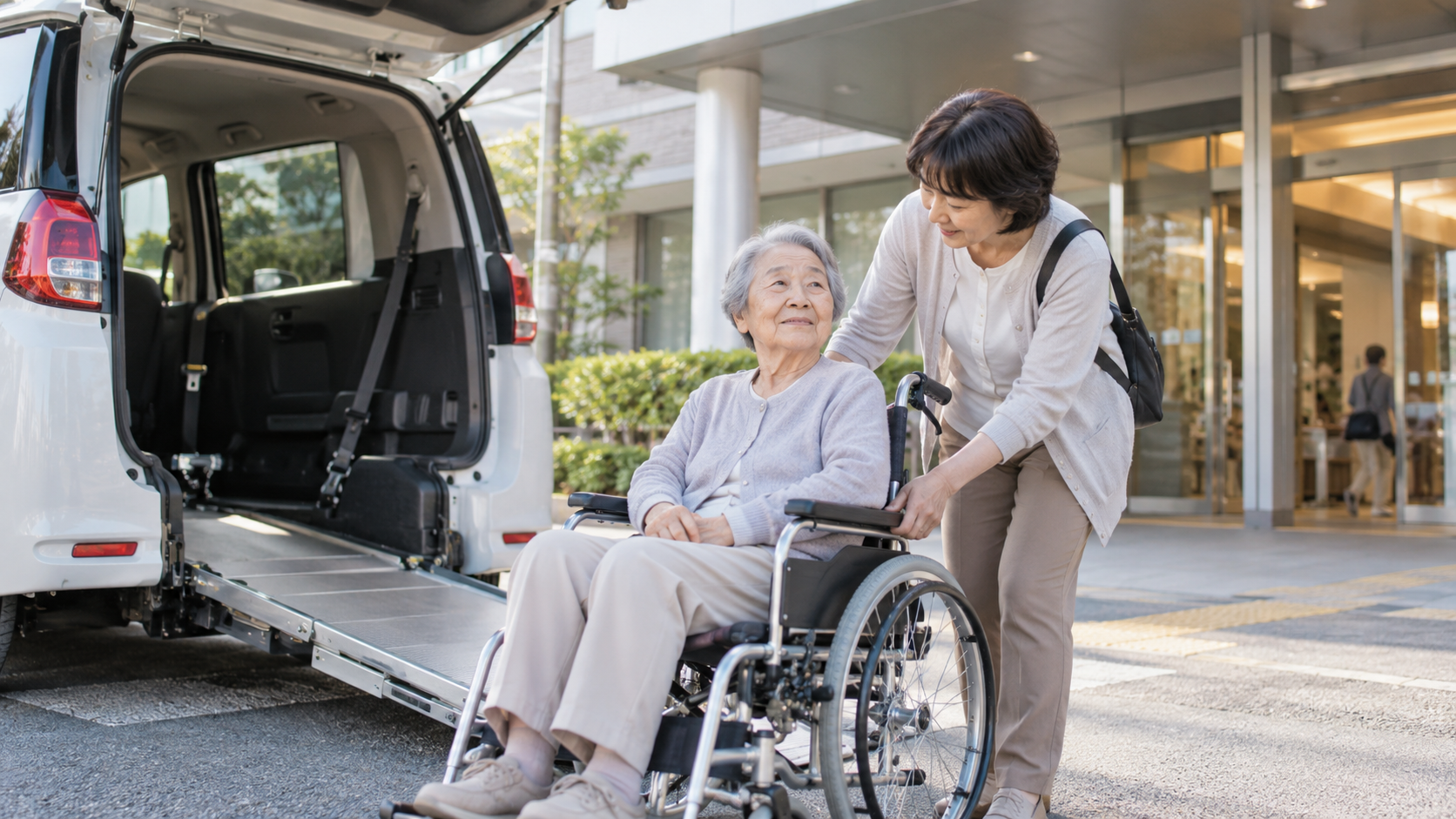 病院前で車椅子に座る高齢の母に娘が付き添い、スロープ付きの送迎車へ乗る準備をしている様子